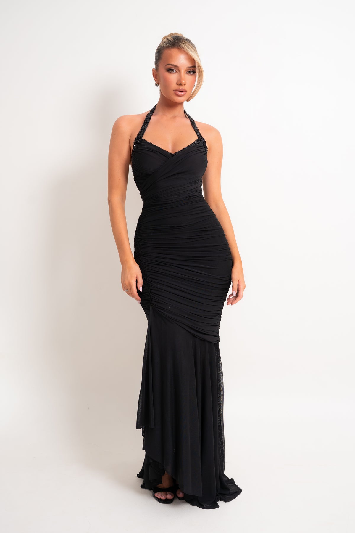 A woman modeling a black halter-neck, ruched mermaid gown against a plain white studio background.