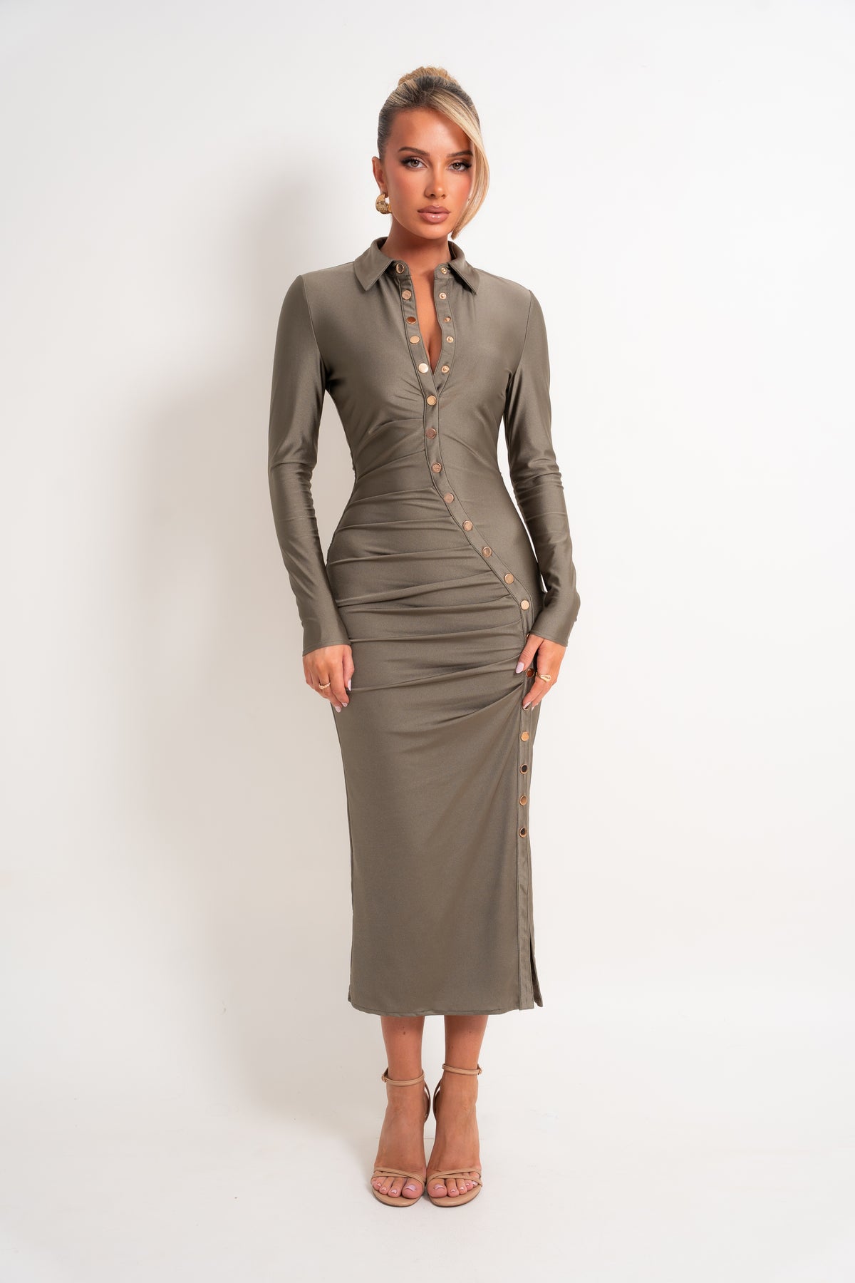 A woman modeling a fitted long-sleeve midi dress in taupe brown with gold-tone buttons down the front and along the side, paired with nude high-heeled sandals against a plain white studio background.