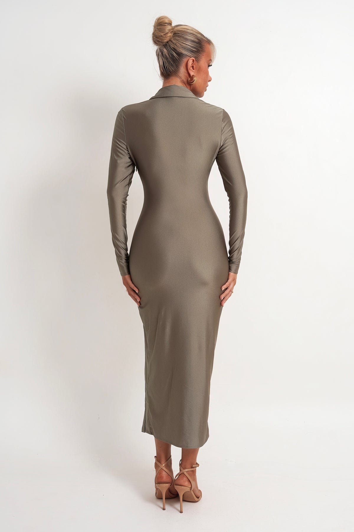 A woman viewed from the back wearing a fitted long-sleeve midi dress in a taupe-brown color, paired with tan high-heeled sandals, against a plain white studio background.