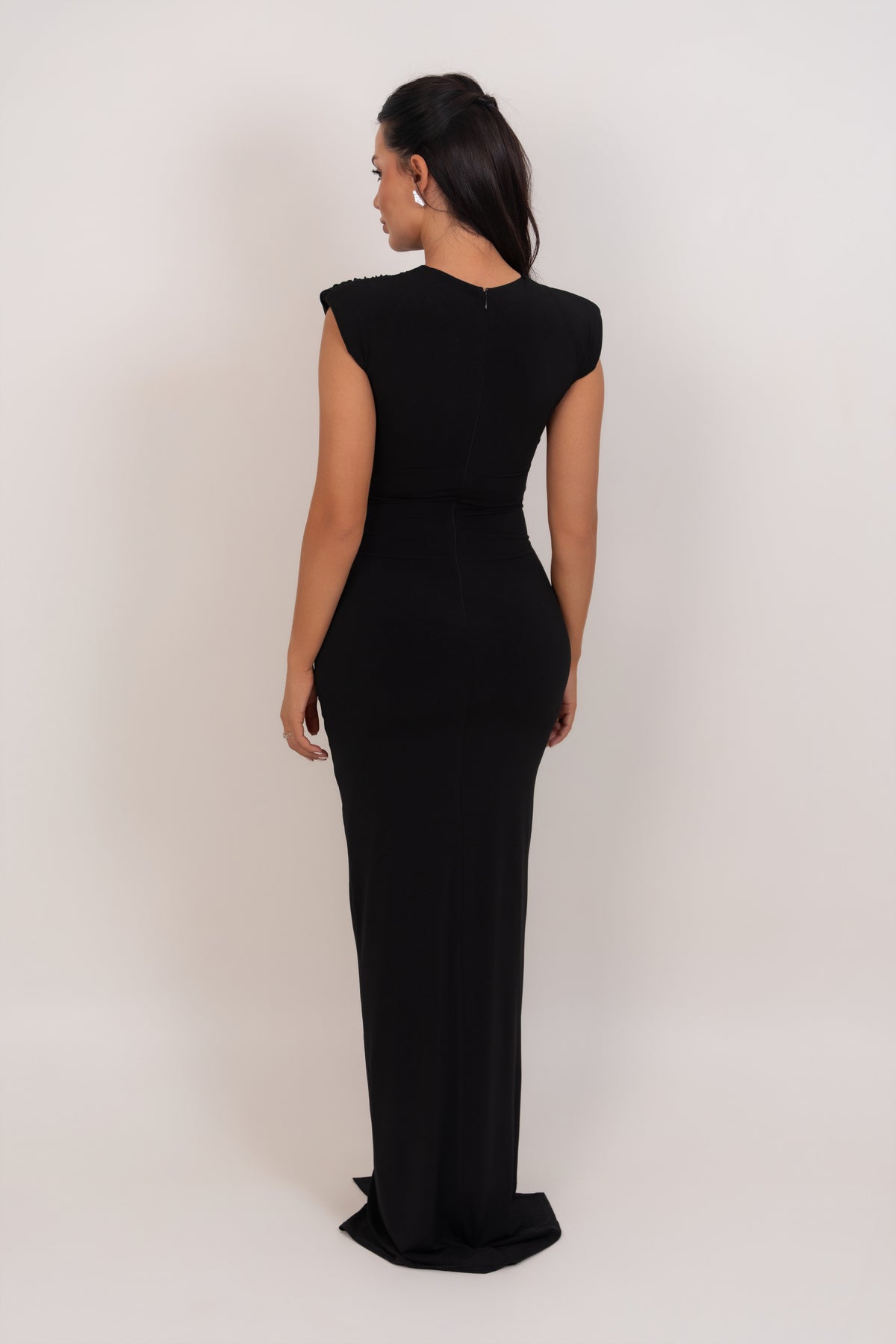 A woman modeling a black sleeveless floor-length gown from the back, standing against a plain light gray studio background.