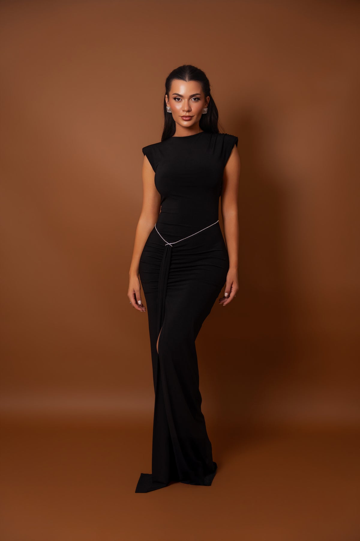 A woman modeling a black sleeveless maxi dress with a high neckline, side slit, and a thin silver chain detail at the waist, set against a solid brown studio backdrop.
