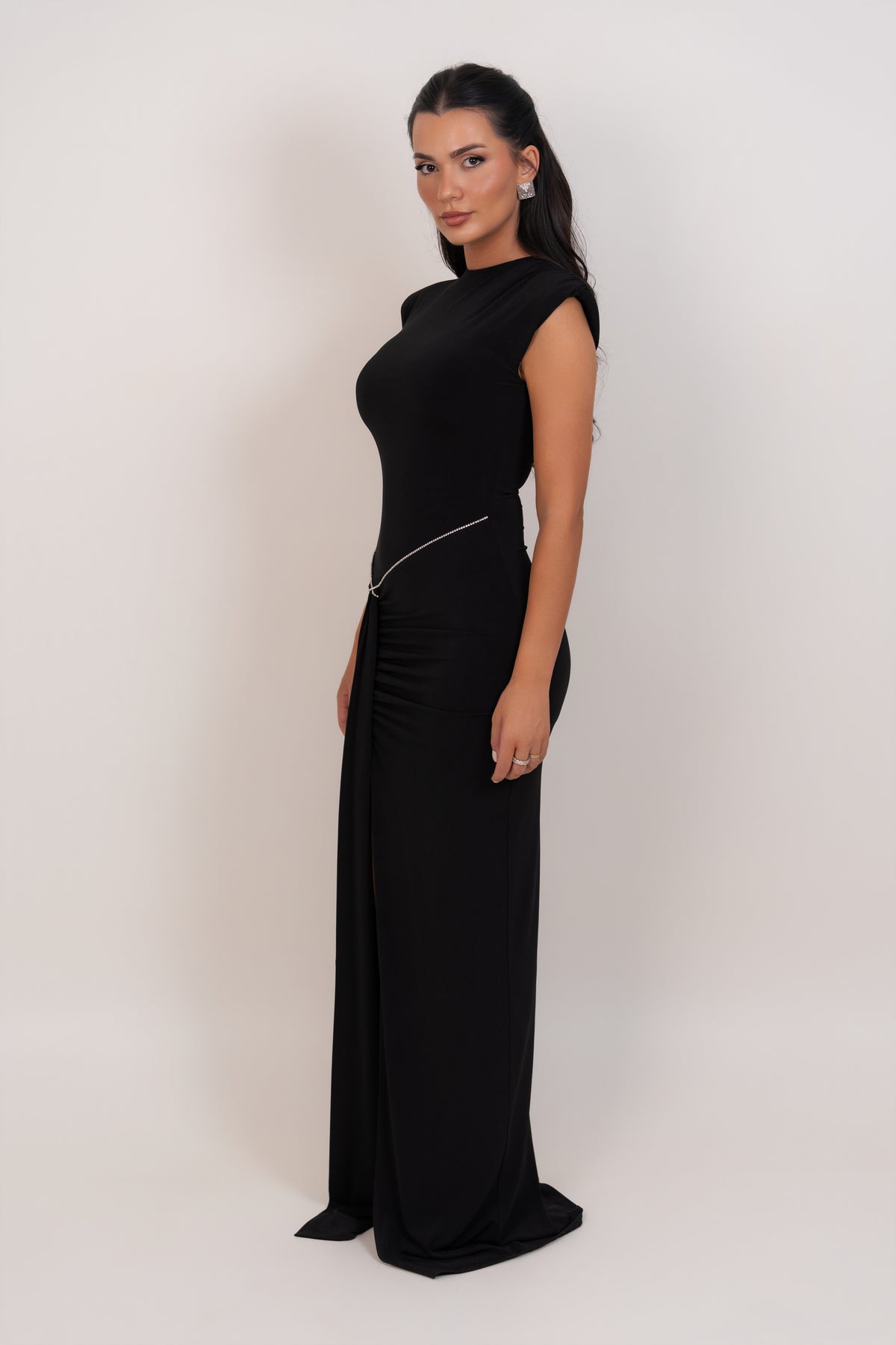 A woman modeling a floor-length black dress with a mock neck, cap sleeves, and a thin silver chain detail at the waist, photographed against a plain light background.