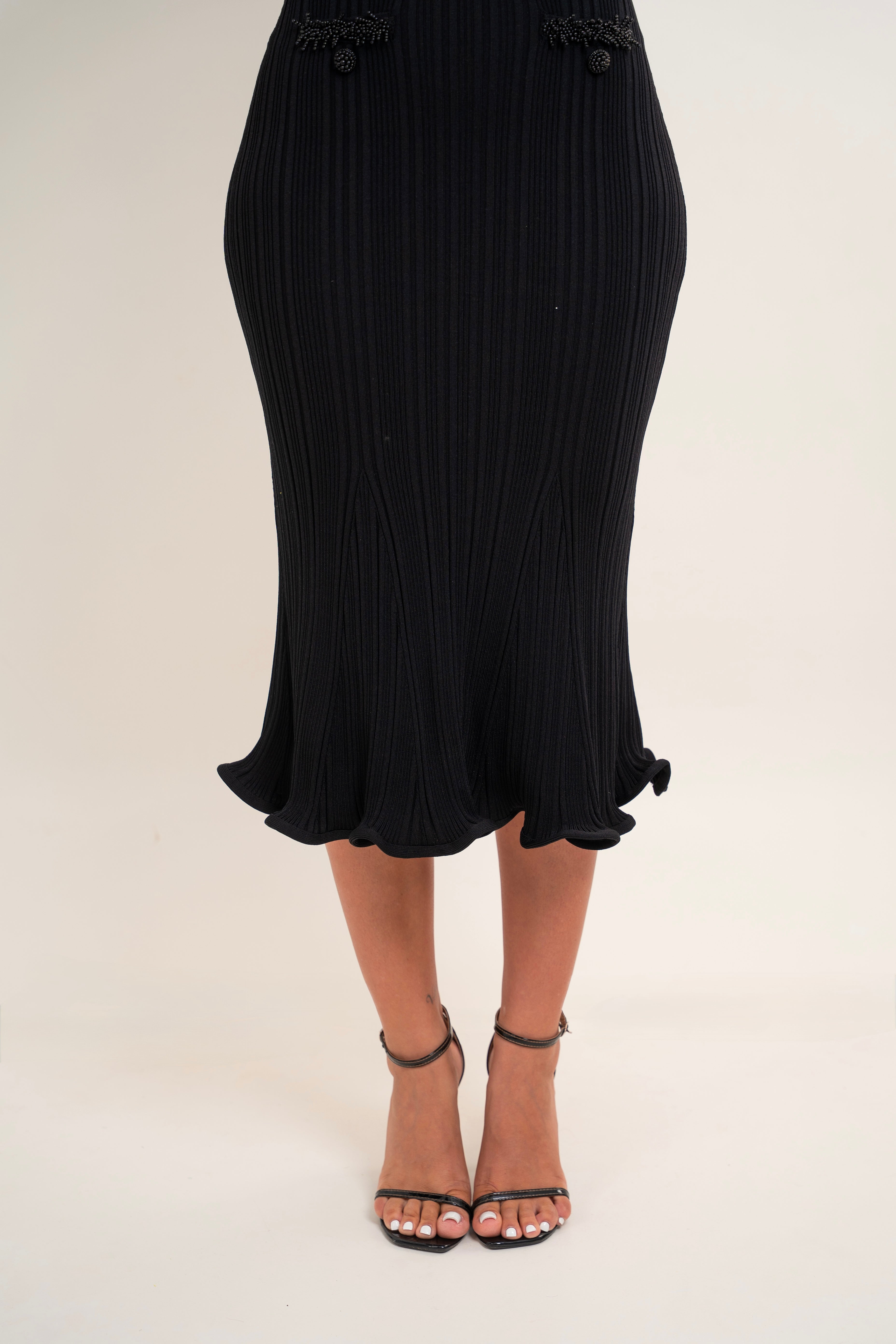 A black pleated midi skirt with a ruffled hem and decorative black bead details at the waist, shown on a model wearing black strappy high-heeled sandals against a light beige studio background.