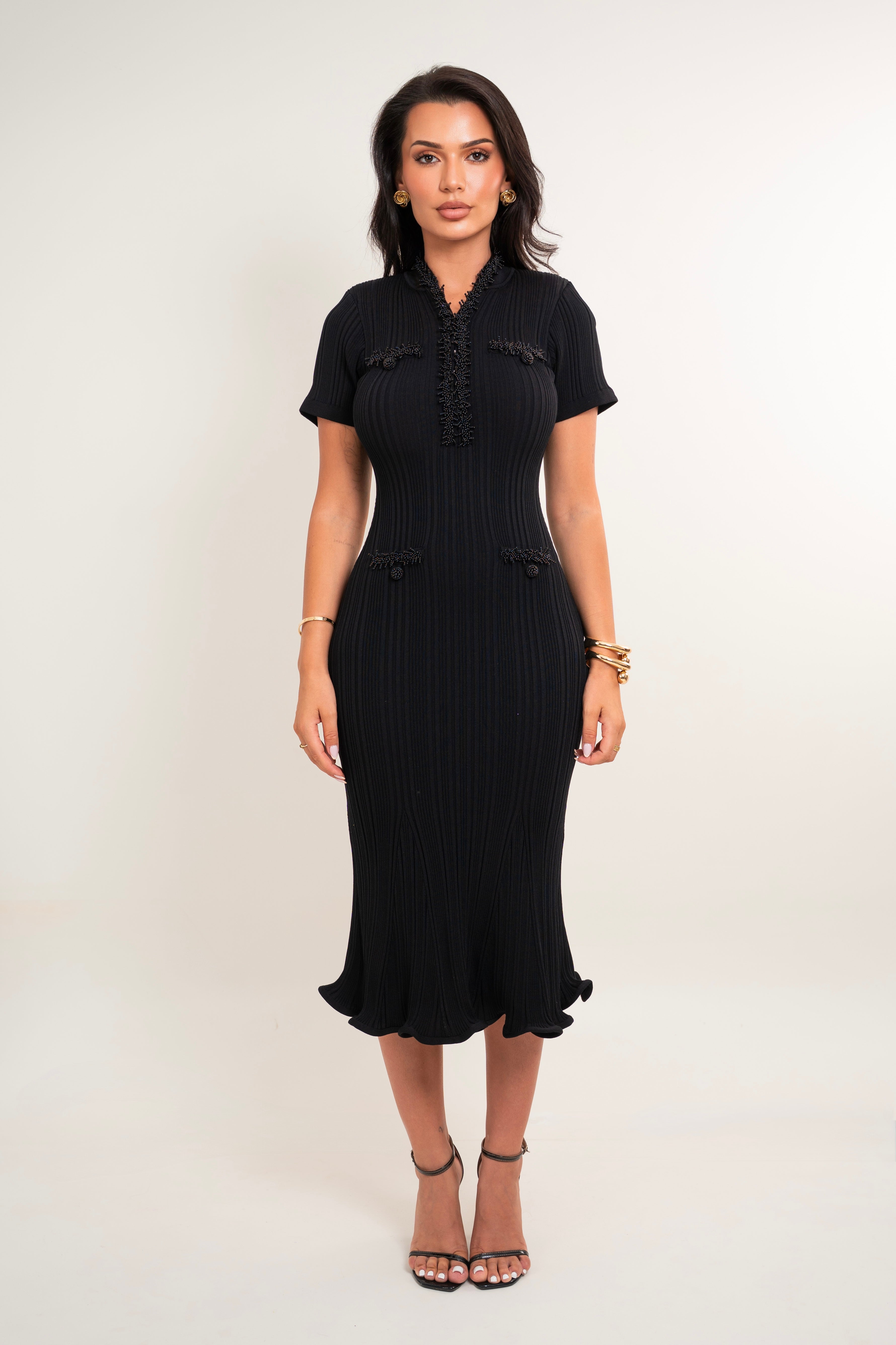A woman modeling a black short-sleeve midi dress with a pleated texture, ruffled collar, and ruffled hem, standing against a plain light background.