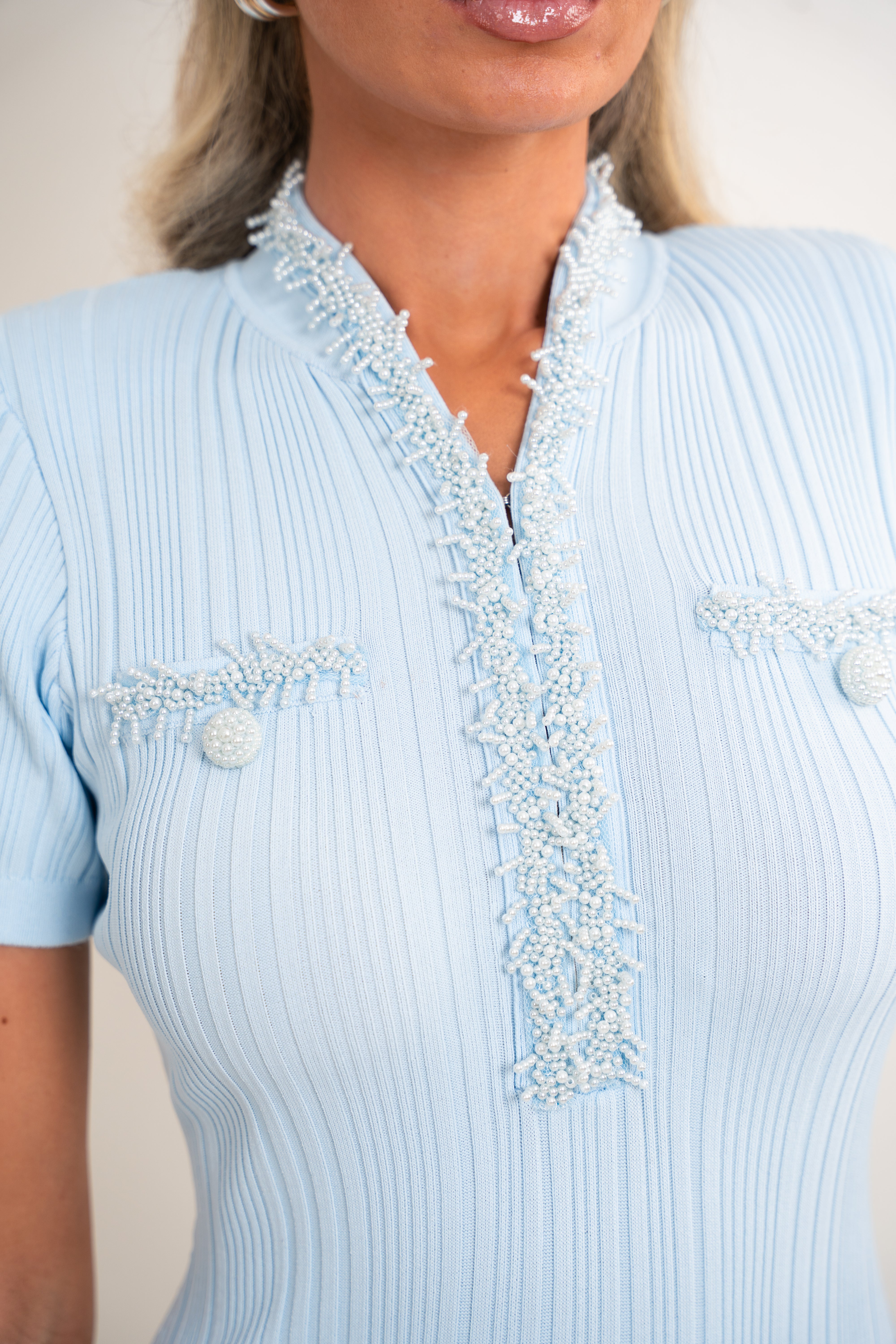 A close-up of a woman wearing a light blue ribbed short-sleeve top with a decorative beaded collar and matching beaded chest details.
