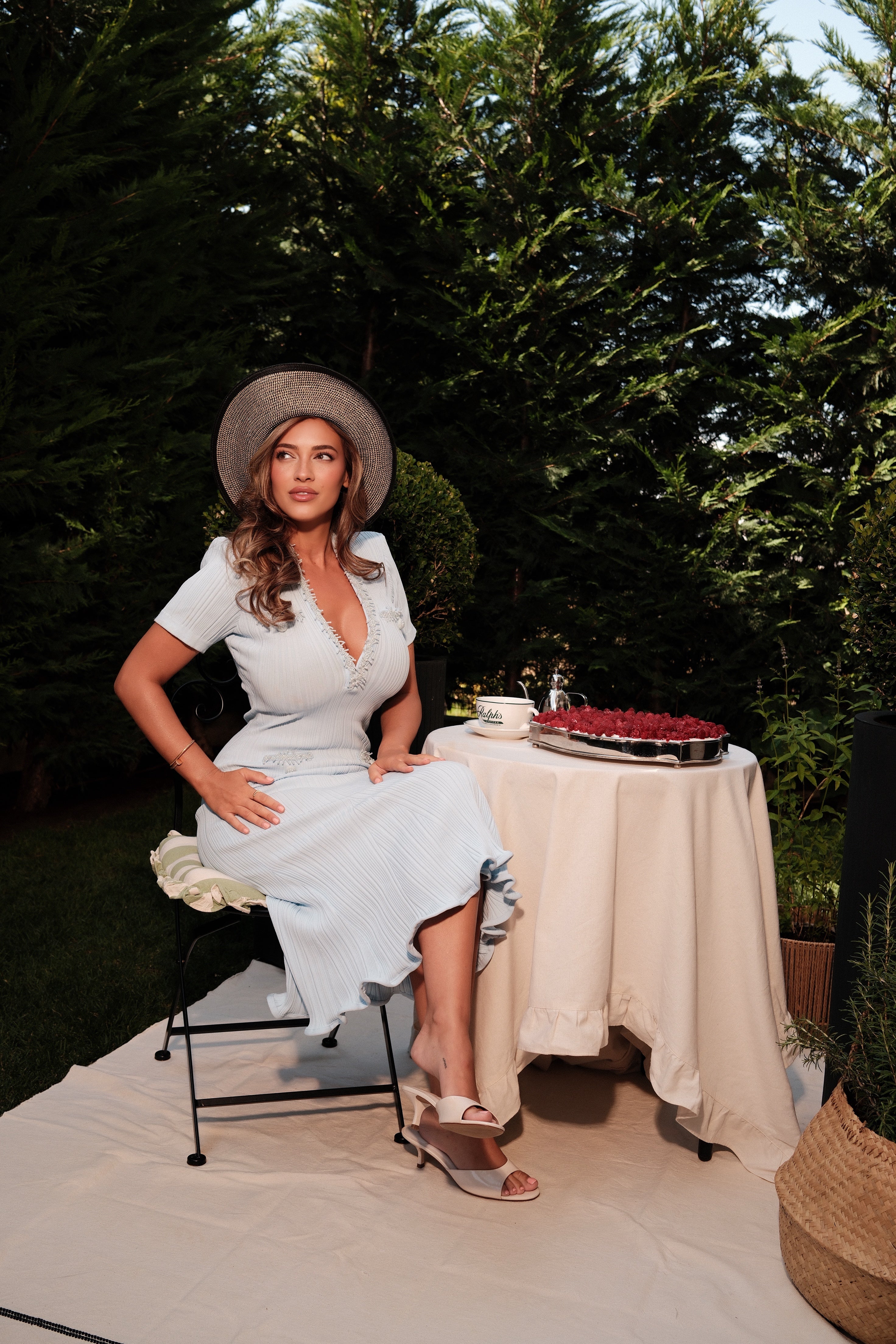 A woman wearing a light blue short-sleeve dress and a wide-brimmed straw hat sits on a chair next to a small table with a white tablecloth in an outdoor garden setting.