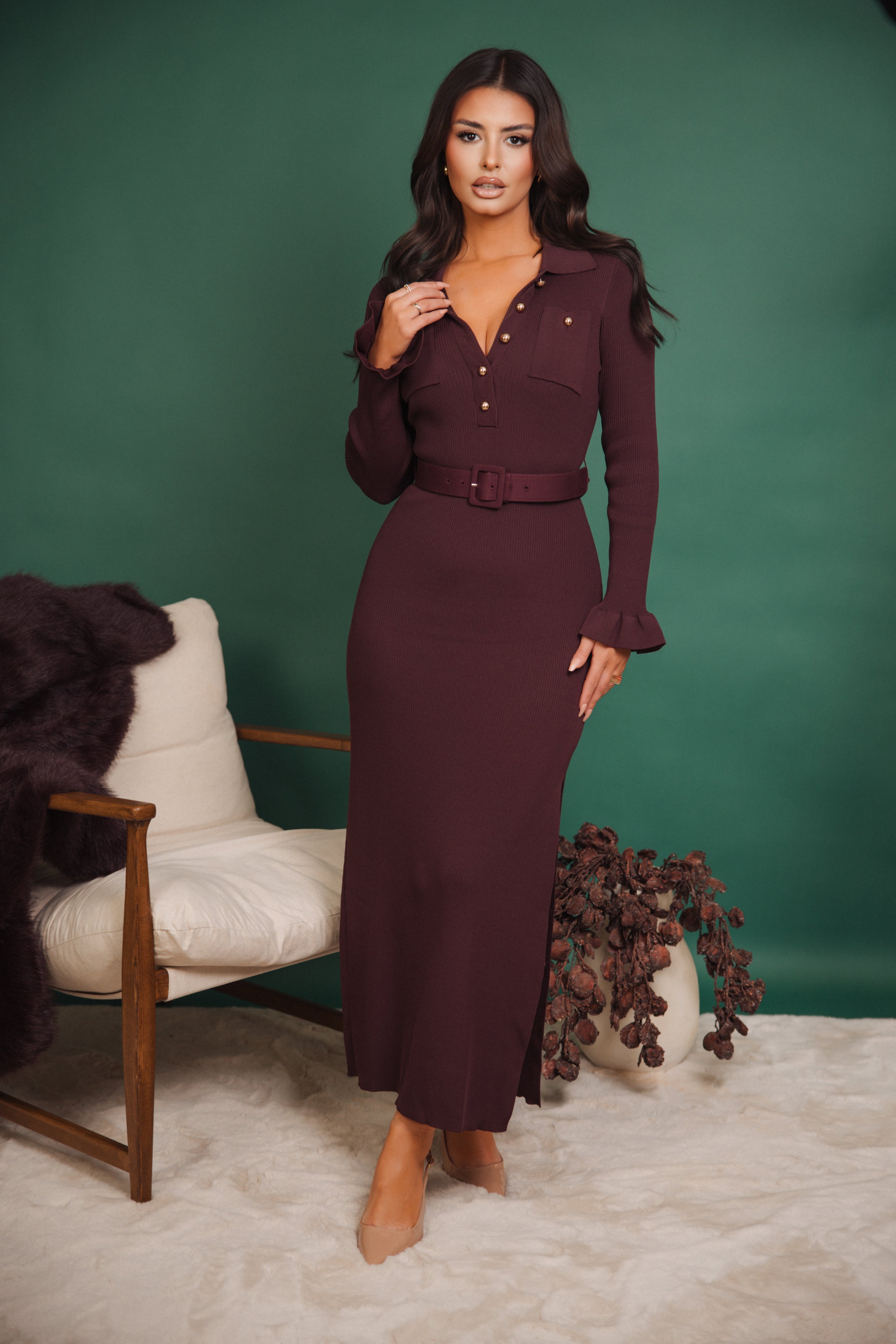 Woman in a burgundy dress standing in front of a green wall with a chair and decorative items.