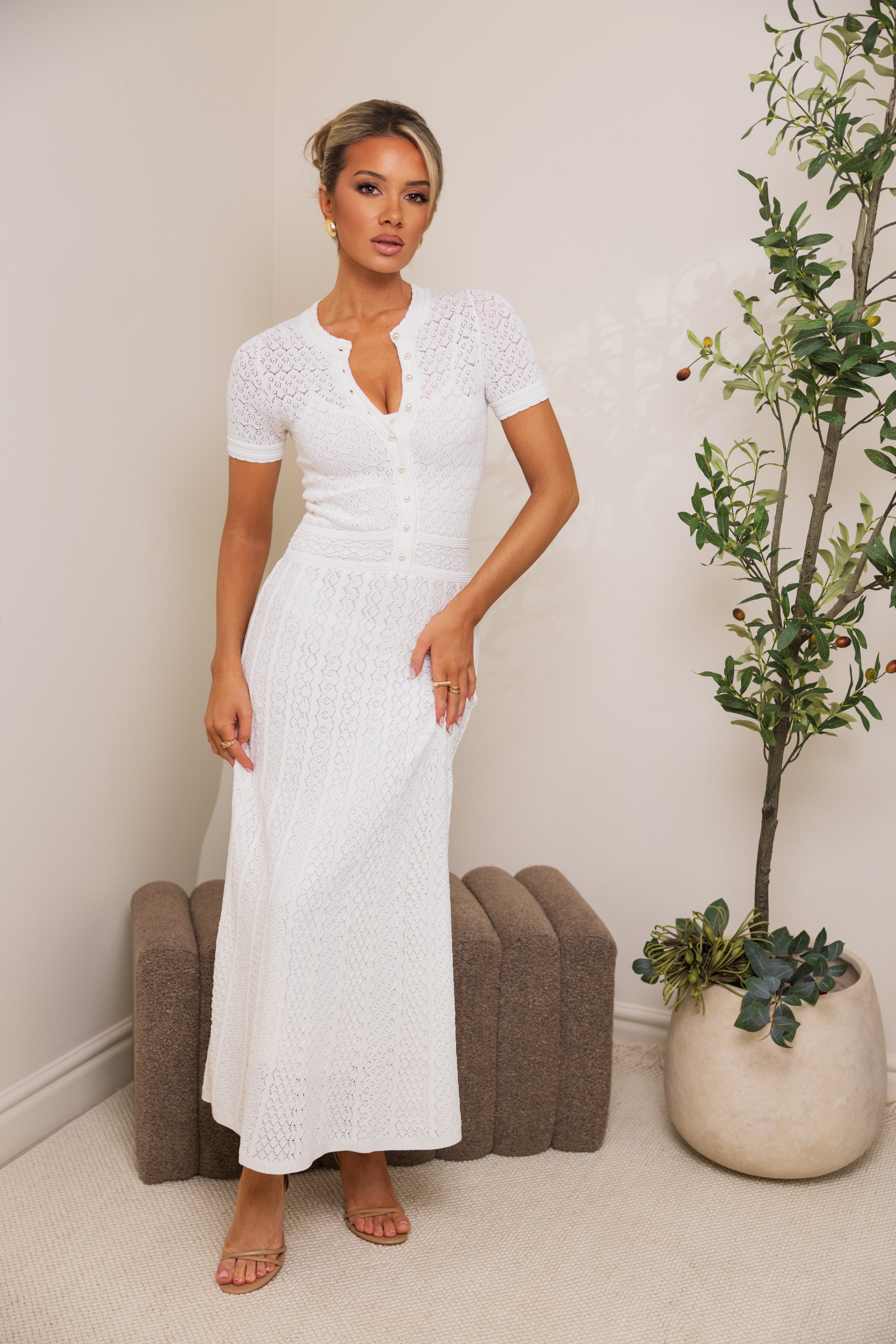 Woman wearing a white lace dress standing in a room with a plant and ottoman.