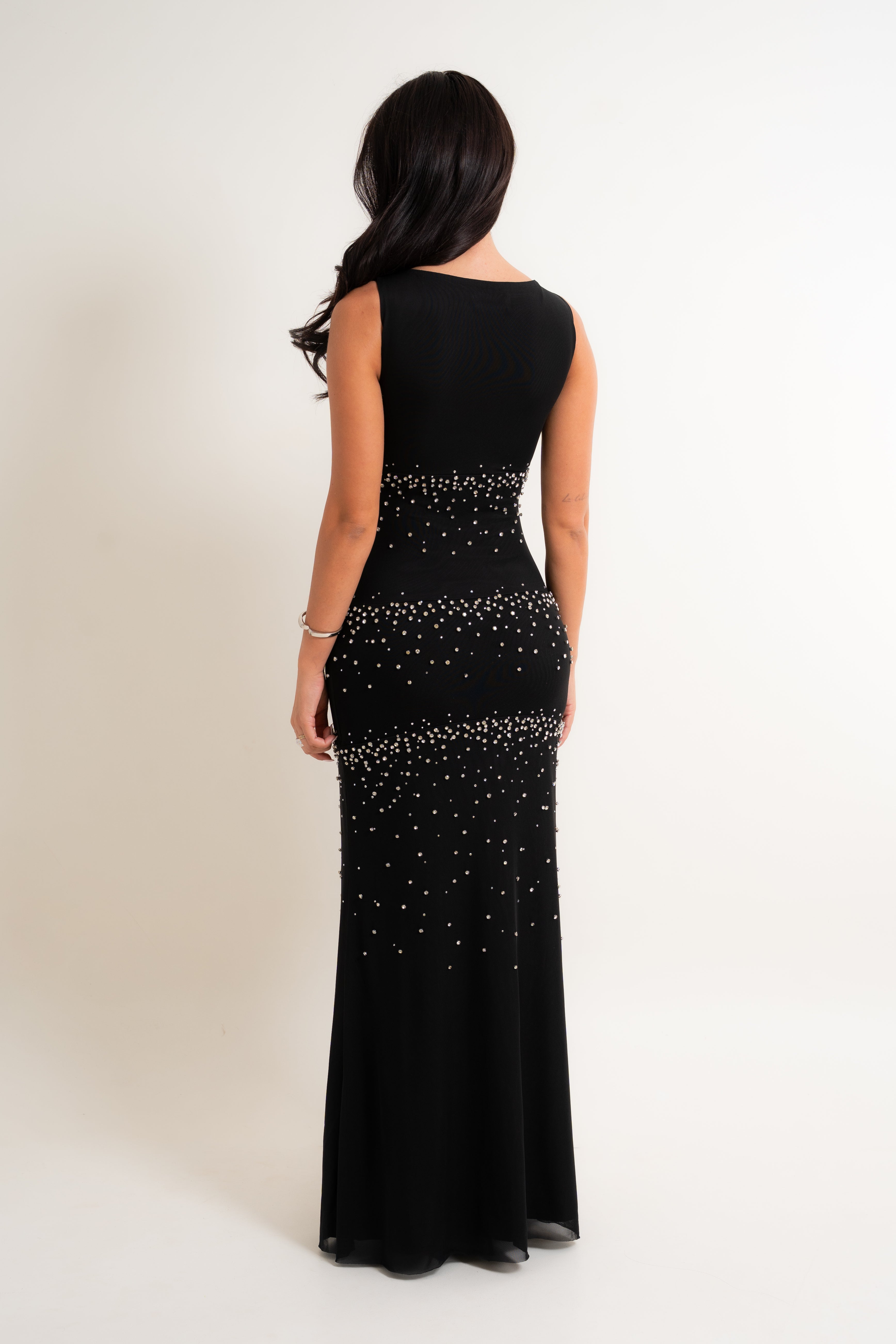A woman viewed from the back wearing a sleeveless black floor-length gown with horizontal rows of sparkling rhinestone embellishments across the bodice and skirt, set against a plain off-white studio background.