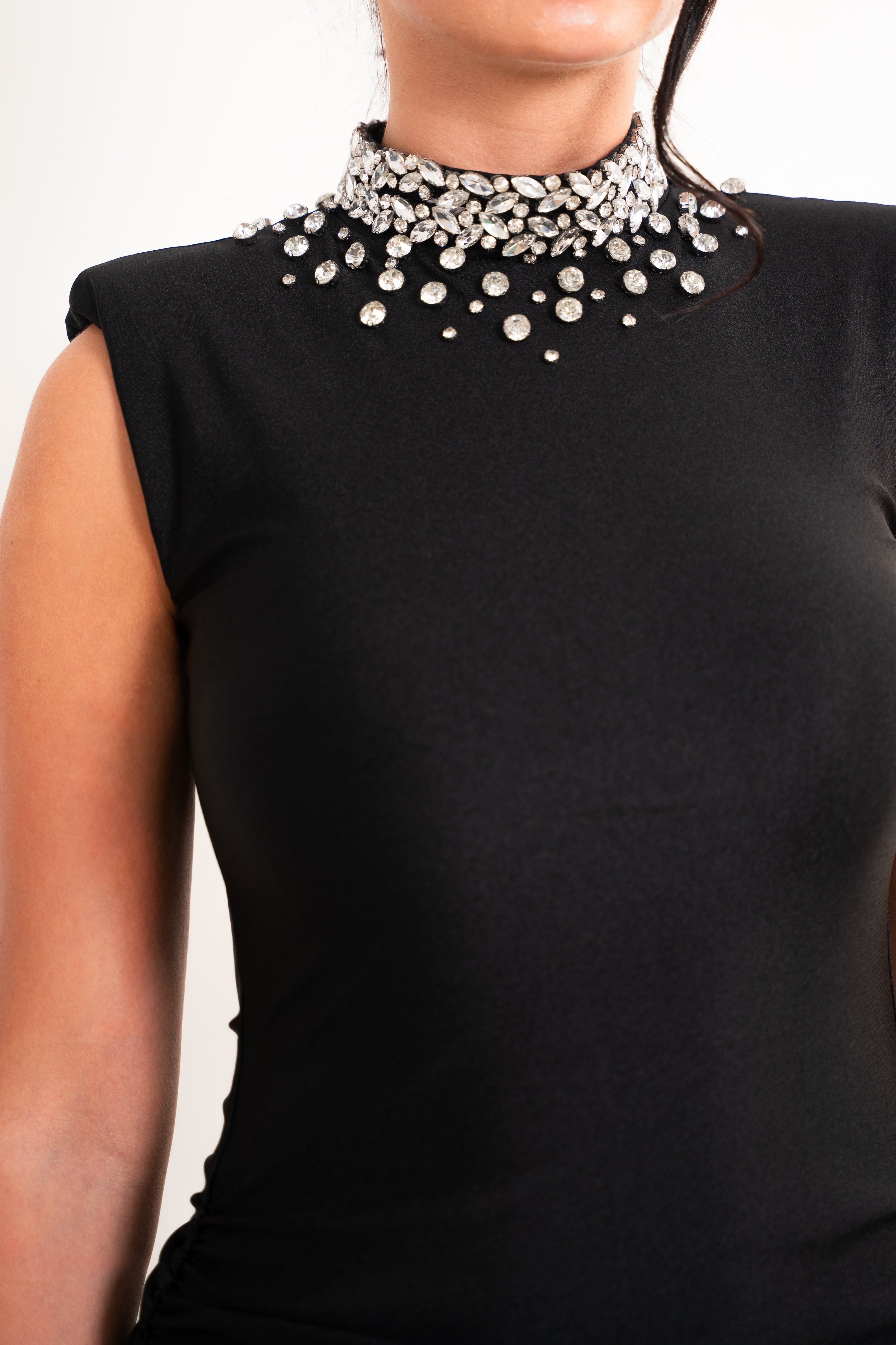 A close-up of a woman wearing a black sleeveless top with a high mock neck heavily embellished with silver rhinestones and clear crystal beads.