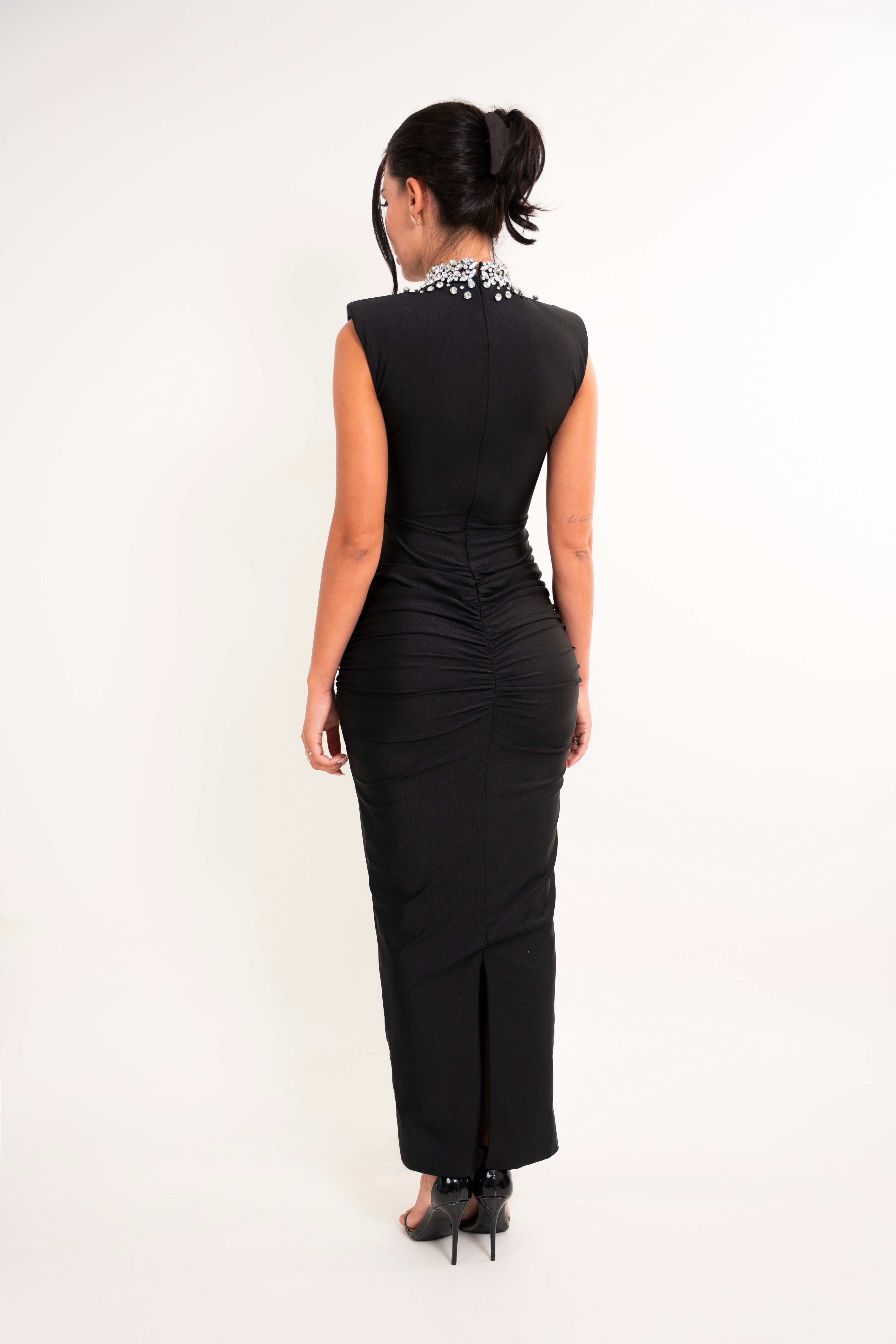 A woman viewed from the back wearing a sleeveless black maxi dress with a beaded high neckline and gathered ruching at the waist, paired with black heels against a plain white studio background.