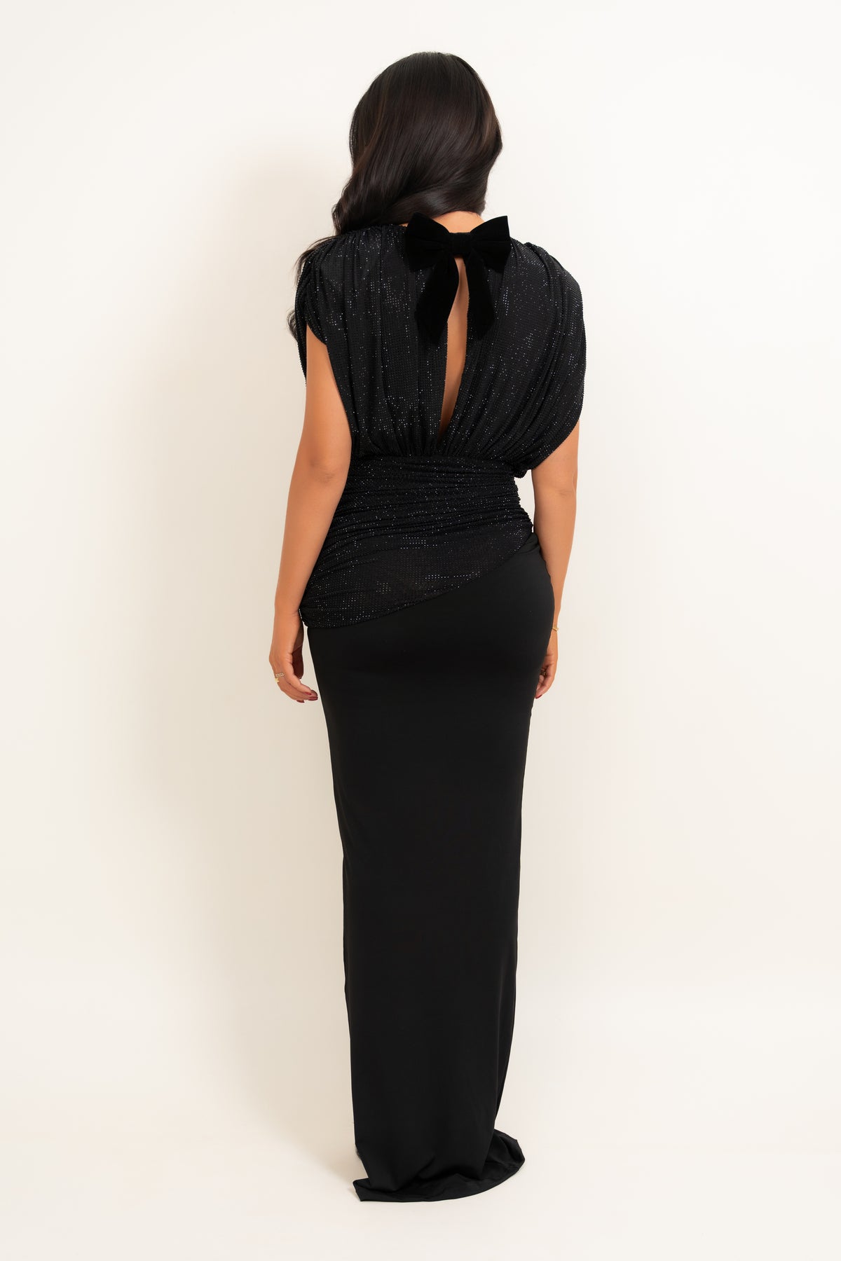 A woman viewed from the back wearing a floor-length black gown with a bow at the back of the neckline, photographed against a plain light studio background.