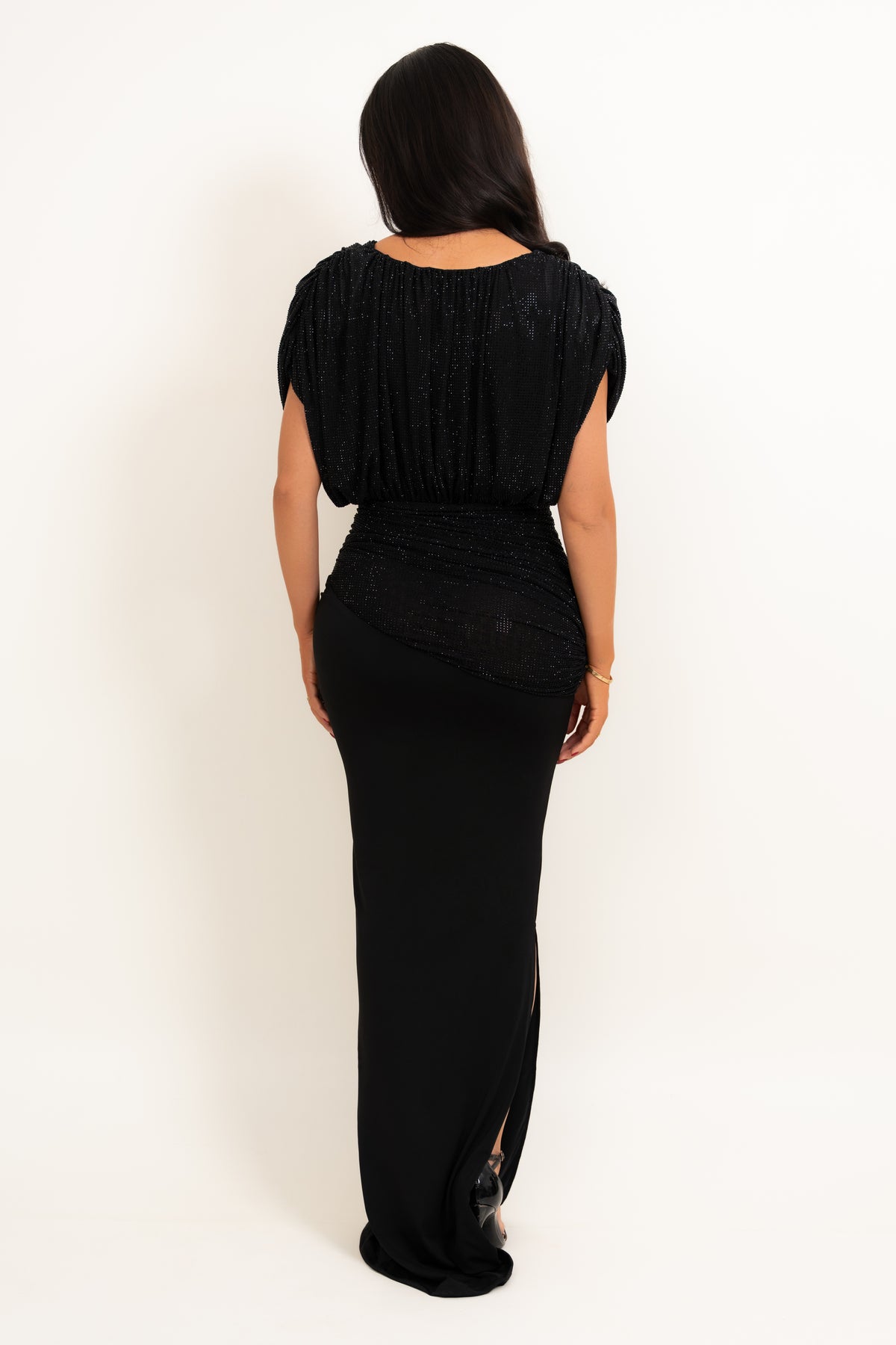A woman viewed from the back wearing a floor-length black gown with a textured pleated bodice, short sleeves, and a fitted skirt with a side slit, against a plain white studio background.