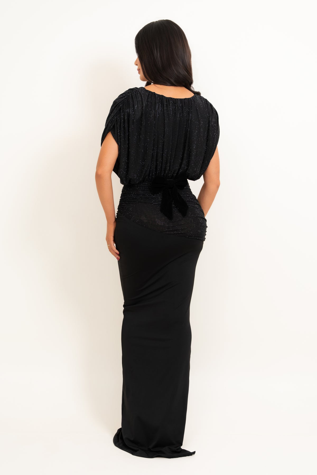 A woman viewed from the back wearing a long black dress with textured pleated short sleeves, a sheer lace-like waist section with a bow, and a fitted floor-length skirt, against a plain light background.