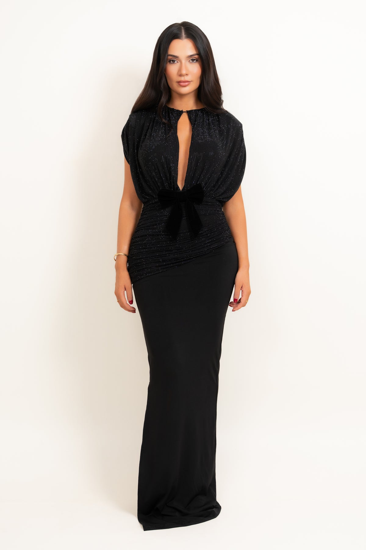A woman modeling a floor-length black gown with a plunging keyhole neckline, short puff sleeves, a gathered waist with a bow detail, and a fitted silhouette, photographed against a plain off-white studio background.