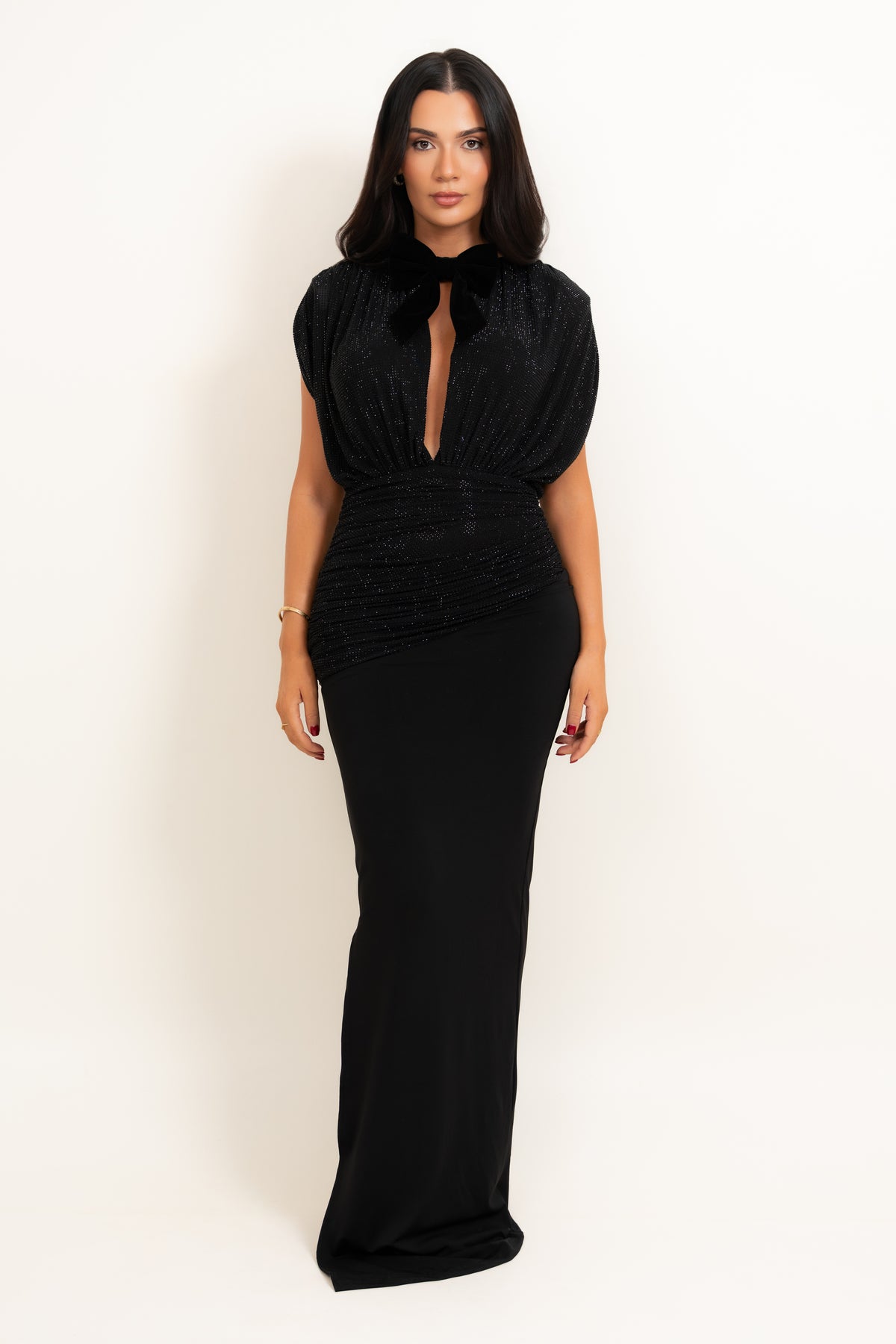 A woman modeling a floor-length black evening gown with a plunging keyhole neckline, sheer cap sleeves, and a fitted silhouette, photographed against a plain off-white studio background.