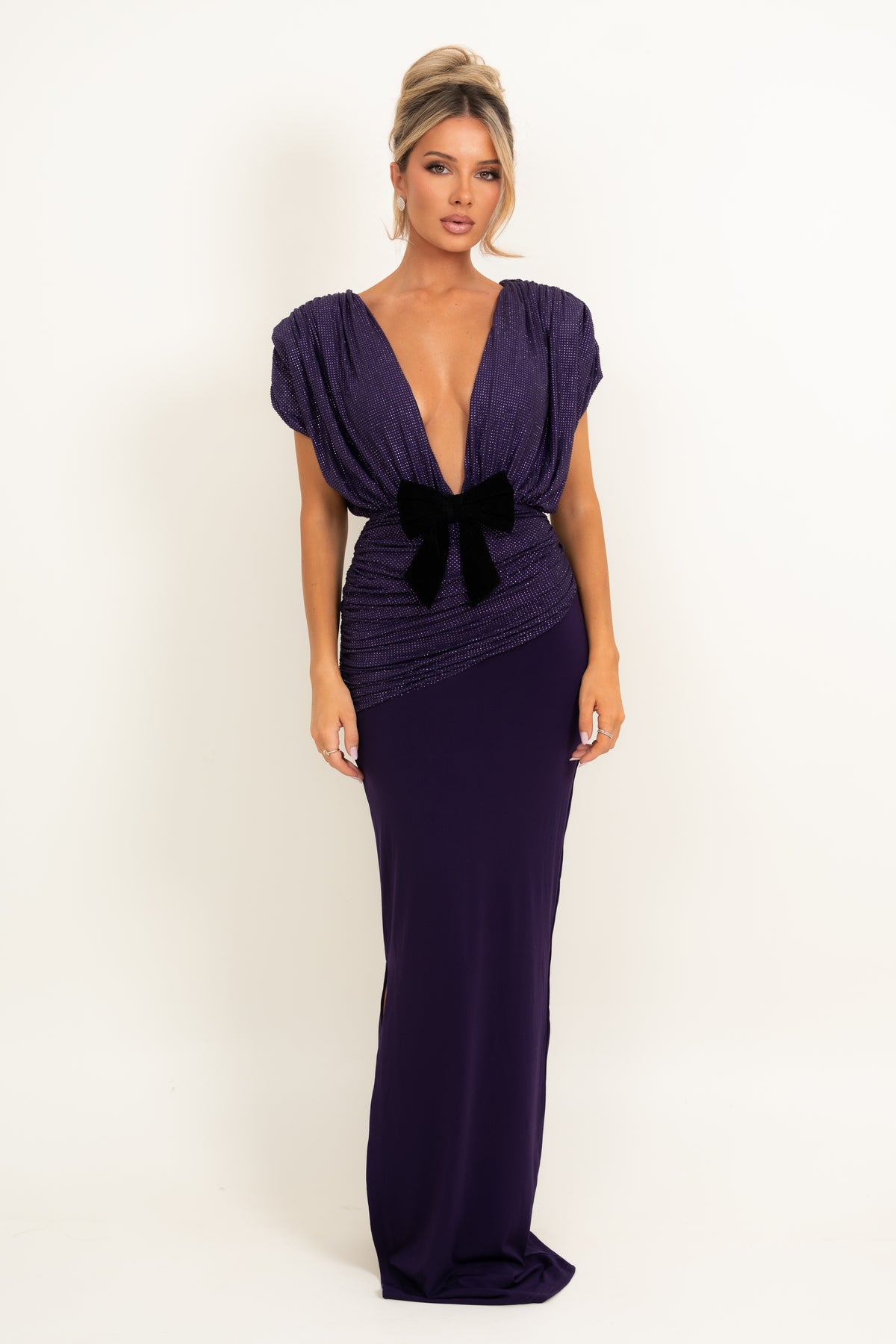 A woman modeling a floor-length deep purple gown with a plunging V-neckline, cap sleeves, ruched detailing, and a black bow at the waist, photographed against a plain light background.