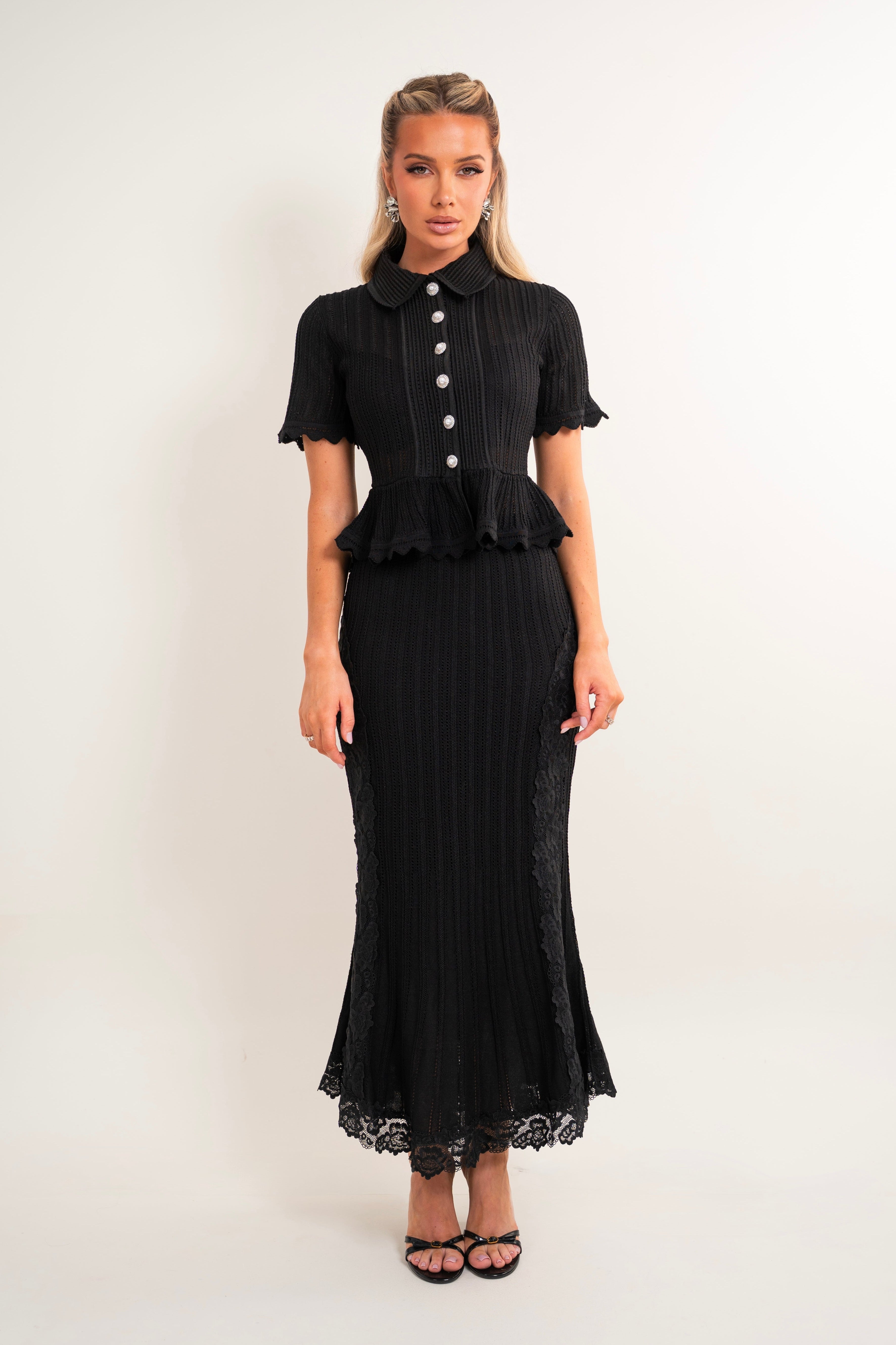 A woman modeling a black short-sleeve midi dress with a Peter Pan collar, buttoned front, peplum waist, and flared lace-trimmed hem, photographed against a plain light background.