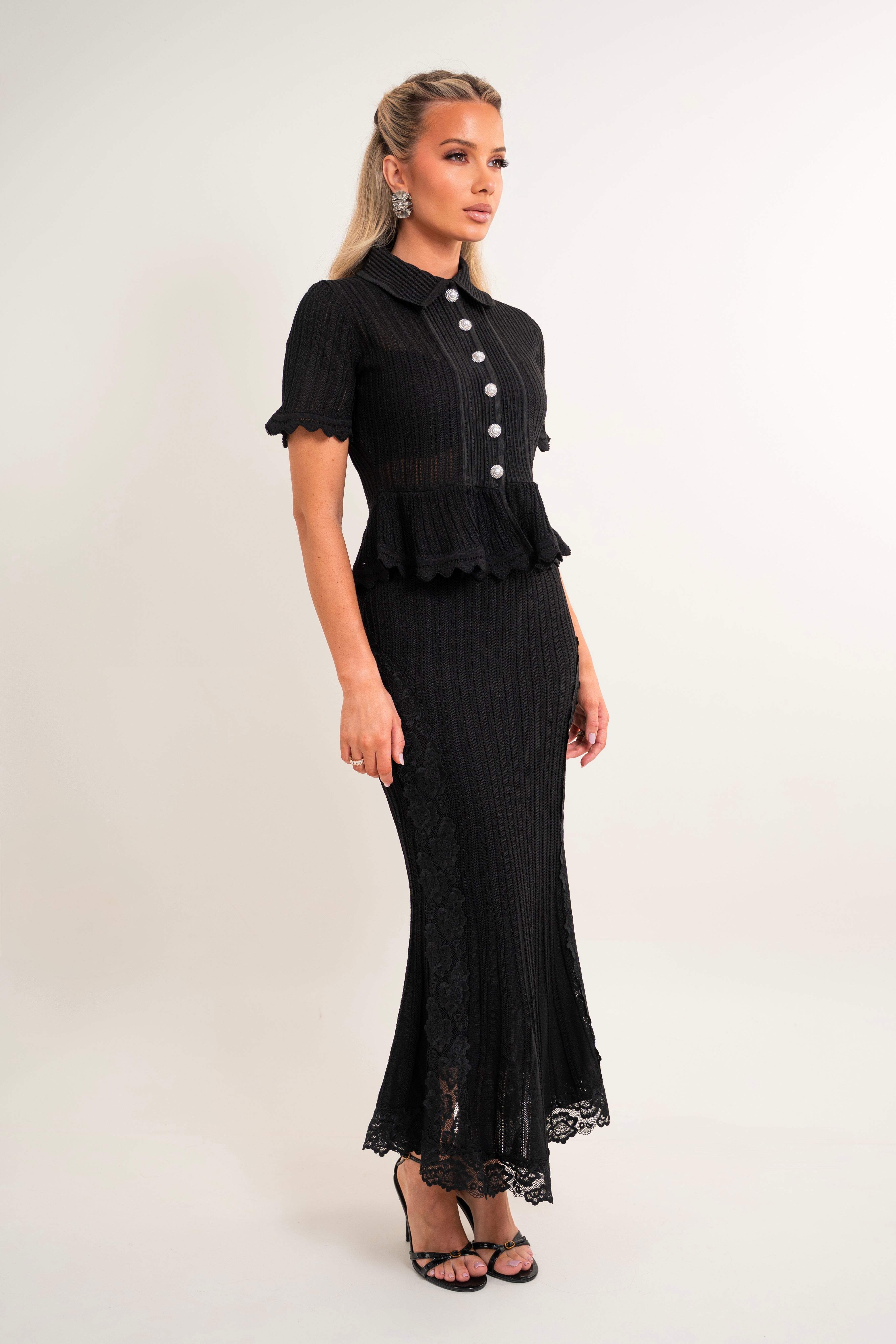 A woman modeling a black short-sleeve midi dress with a ruffled peplum waist, buttoned front, and flared skirt with lace trim, photographed against a plain light studio background.