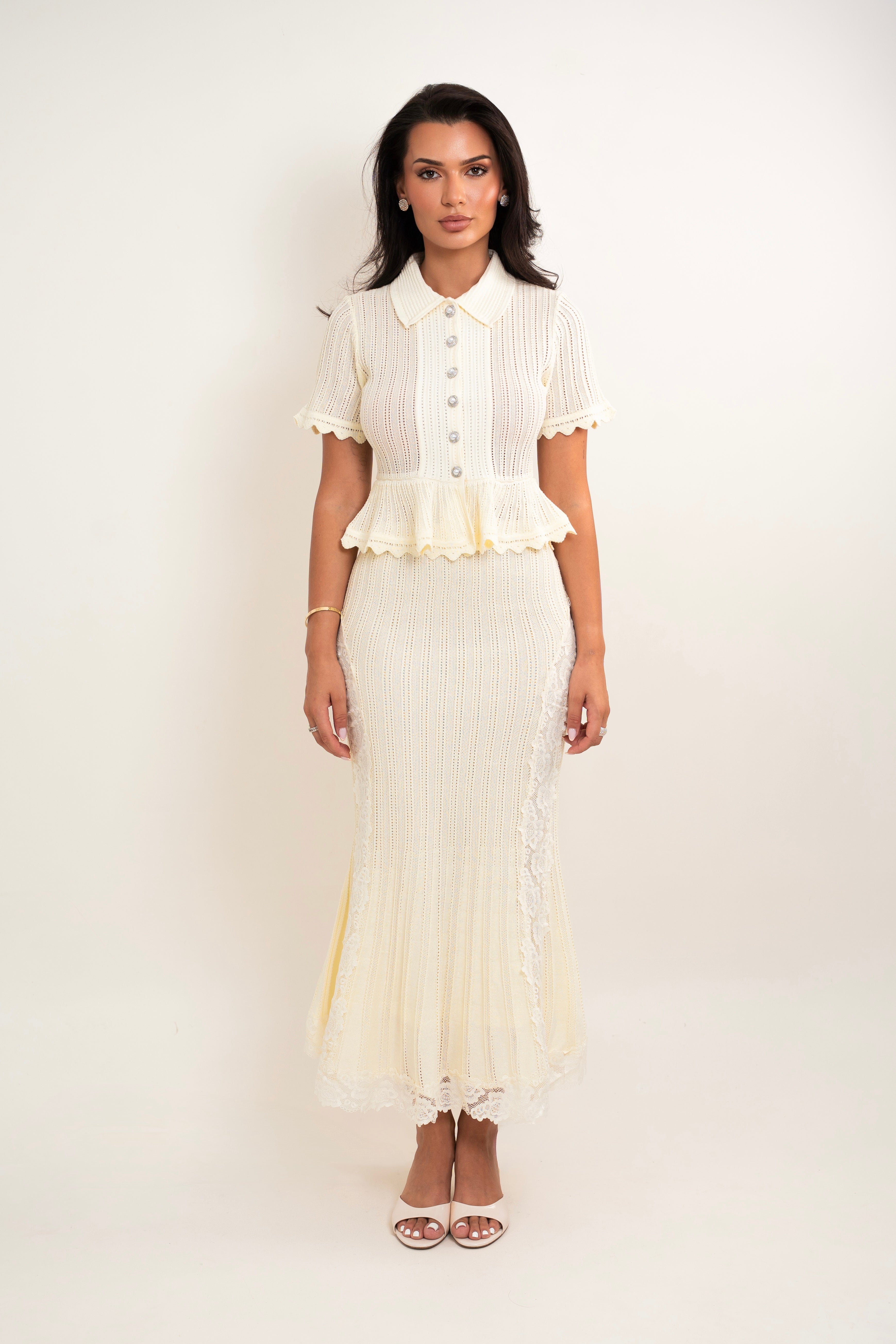 A woman modeling a cream-colored two-piece set consisting of a short-sleeve button-up blouse with a ruffled peplum hem and a matching pleated maxi skirt with lace trim, set against a plain off-white studio background.