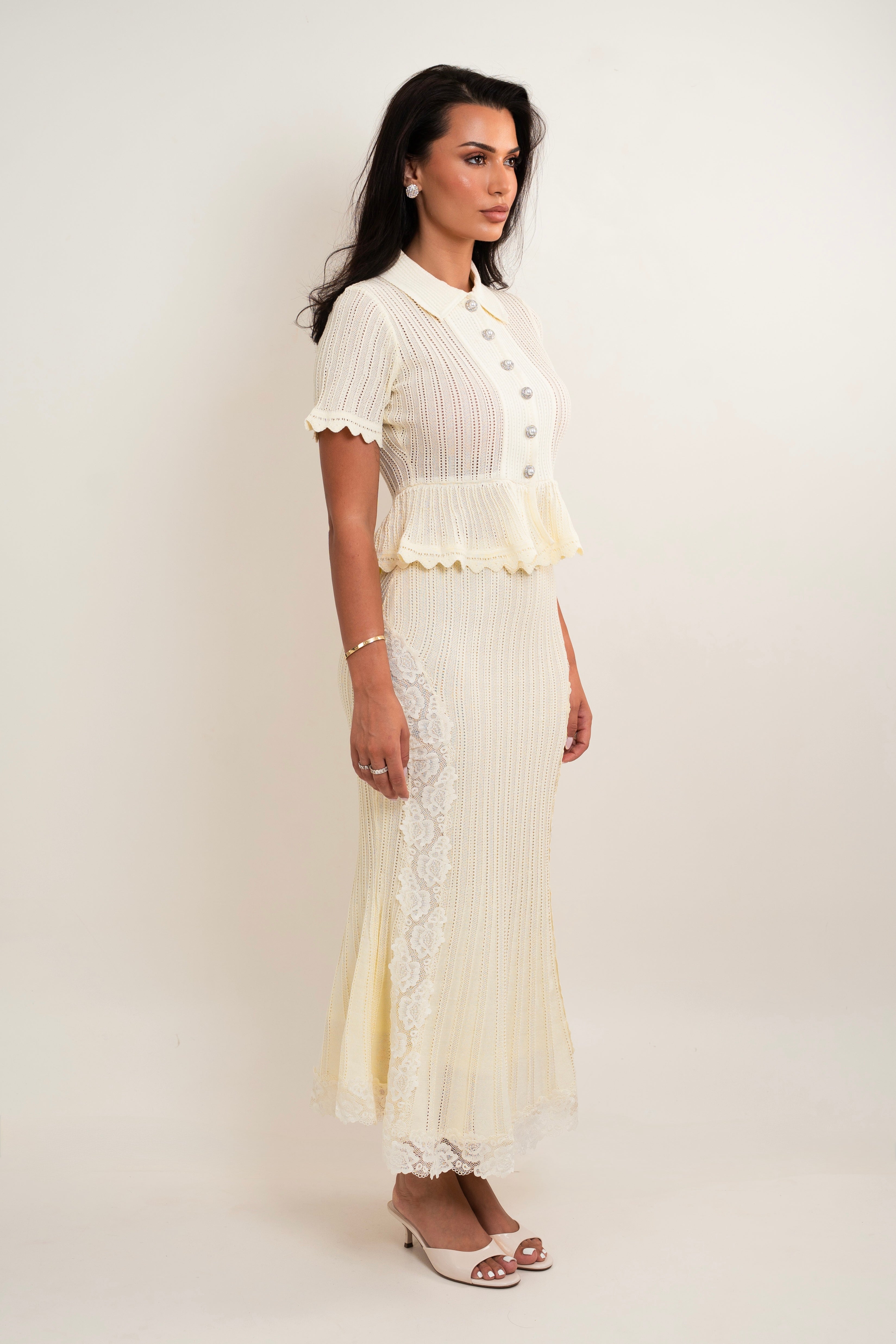 A woman modeling a cream-colored two-piece knit set consisting of a short-sleeve button-front top with a ruffled hem and a matching pleated maxi skirt with lace trim, set against a plain light beige studio background.
