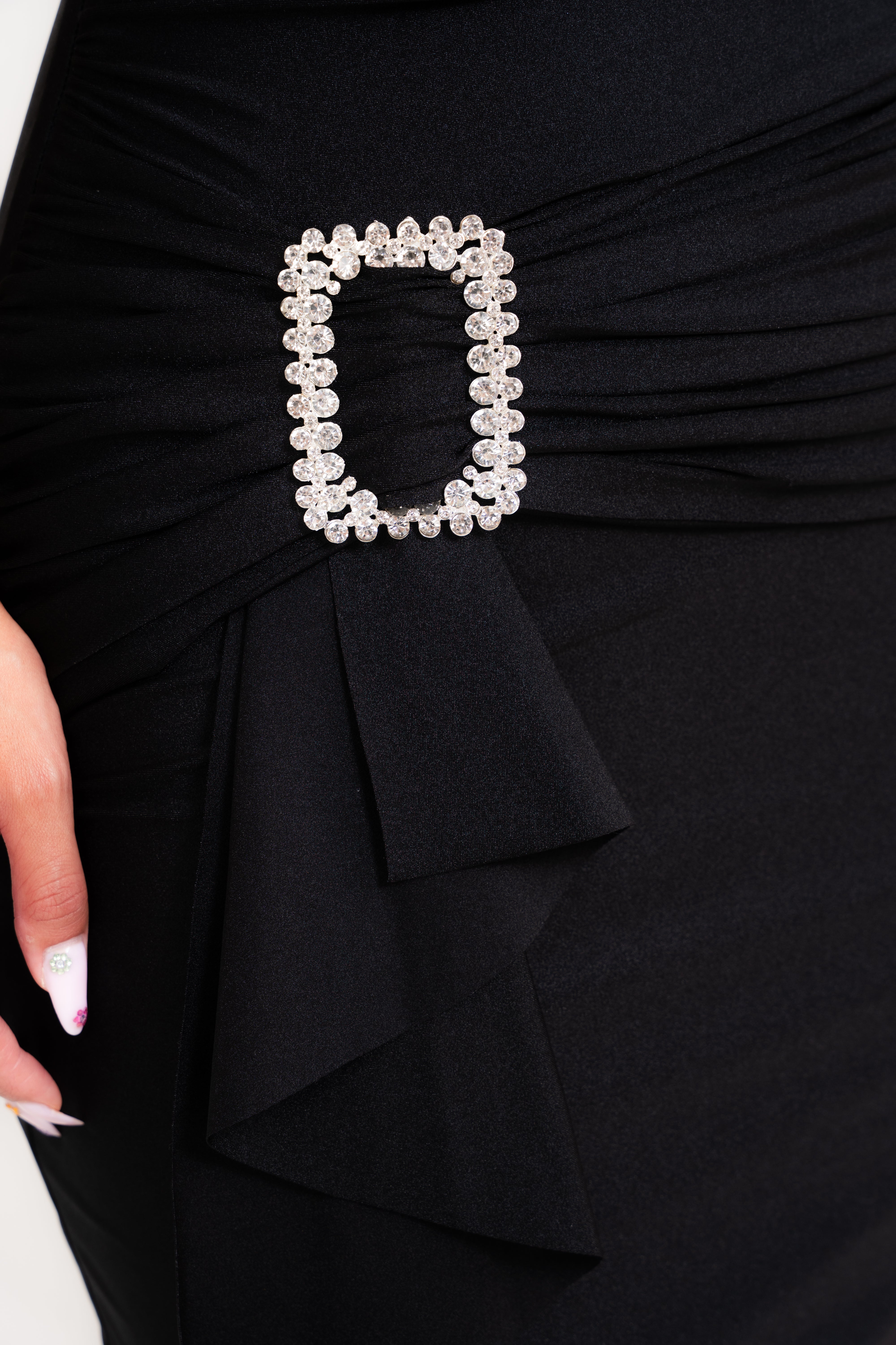 Close-up of a black ruched dress with a decorative rectangular silver rhinestone belt buckle at the waist, with a hand partially visible on the left.
