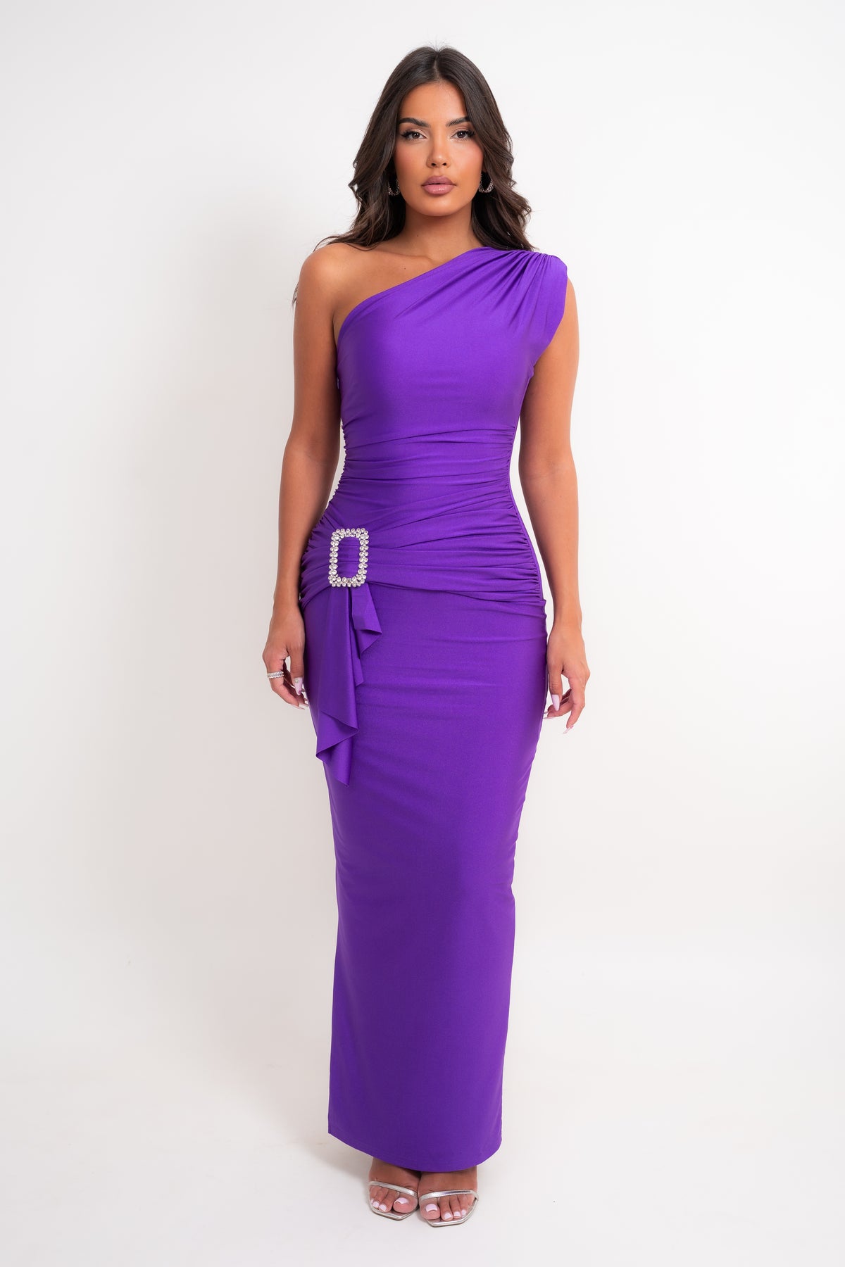 A woman modeling a vibrant purple one-shoulder maxi dress with ruching, a side tie detail, and a rhinestone-embellished belt buckle, set against a plain white studio background.