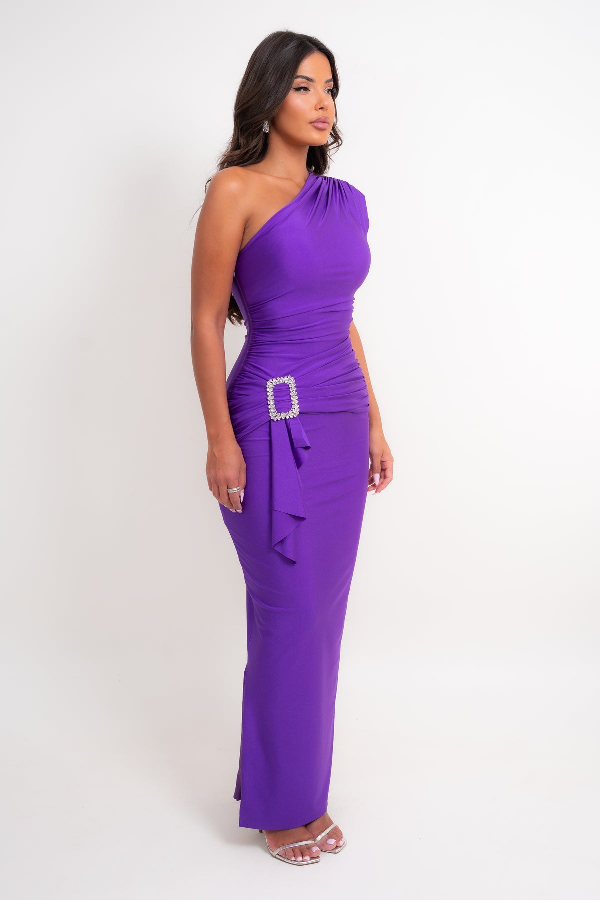 A woman modeling a vibrant purple one-shoulder maxi dress with a ruched bodice, side ruffle, and a silver rhinestone-embellished belt buckle, set against a plain white studio background.