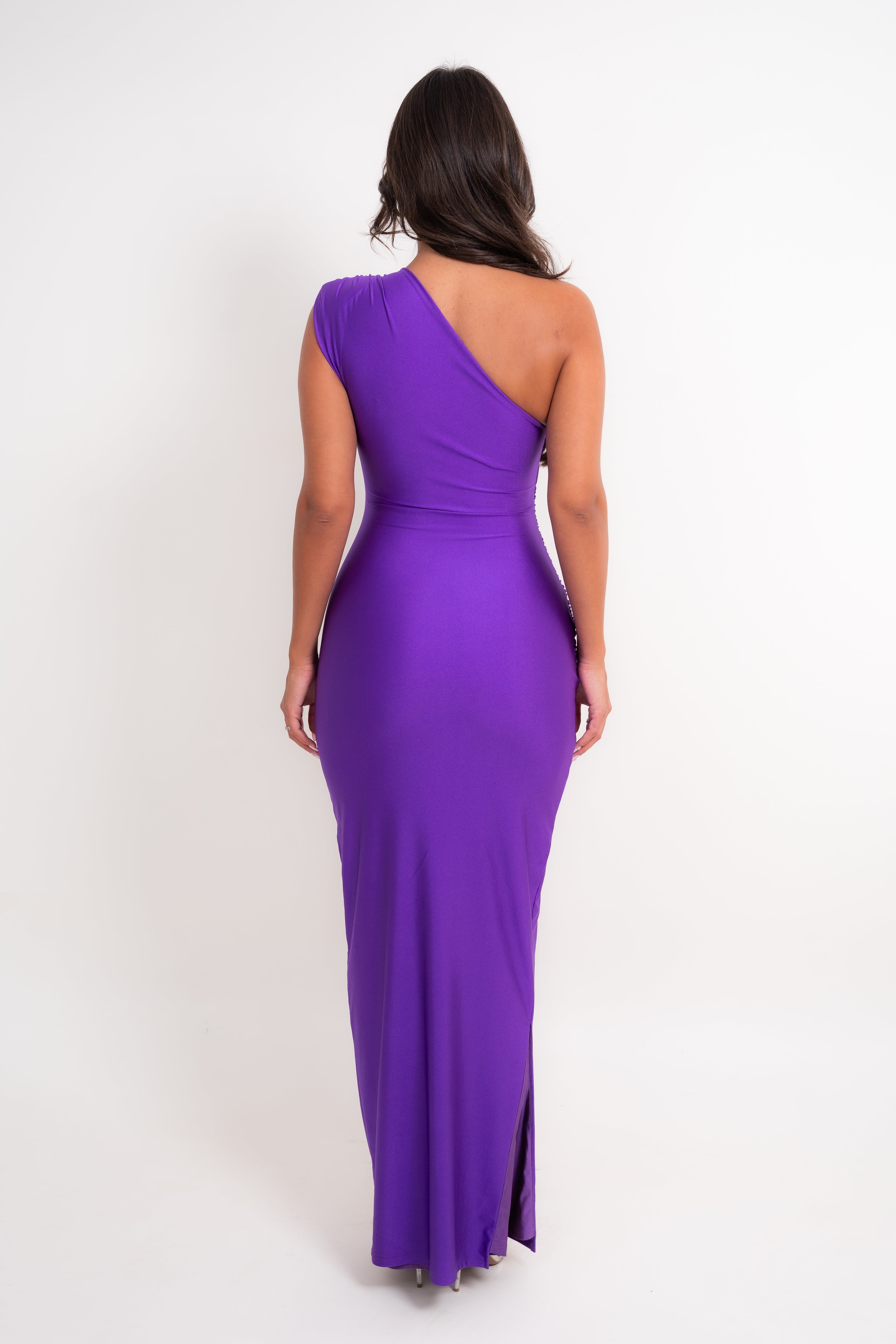 A woman viewed from the back wearing a vibrant purple one-shoulder maxi dress with a fitted waist and a side slit, set against a plain white studio background.