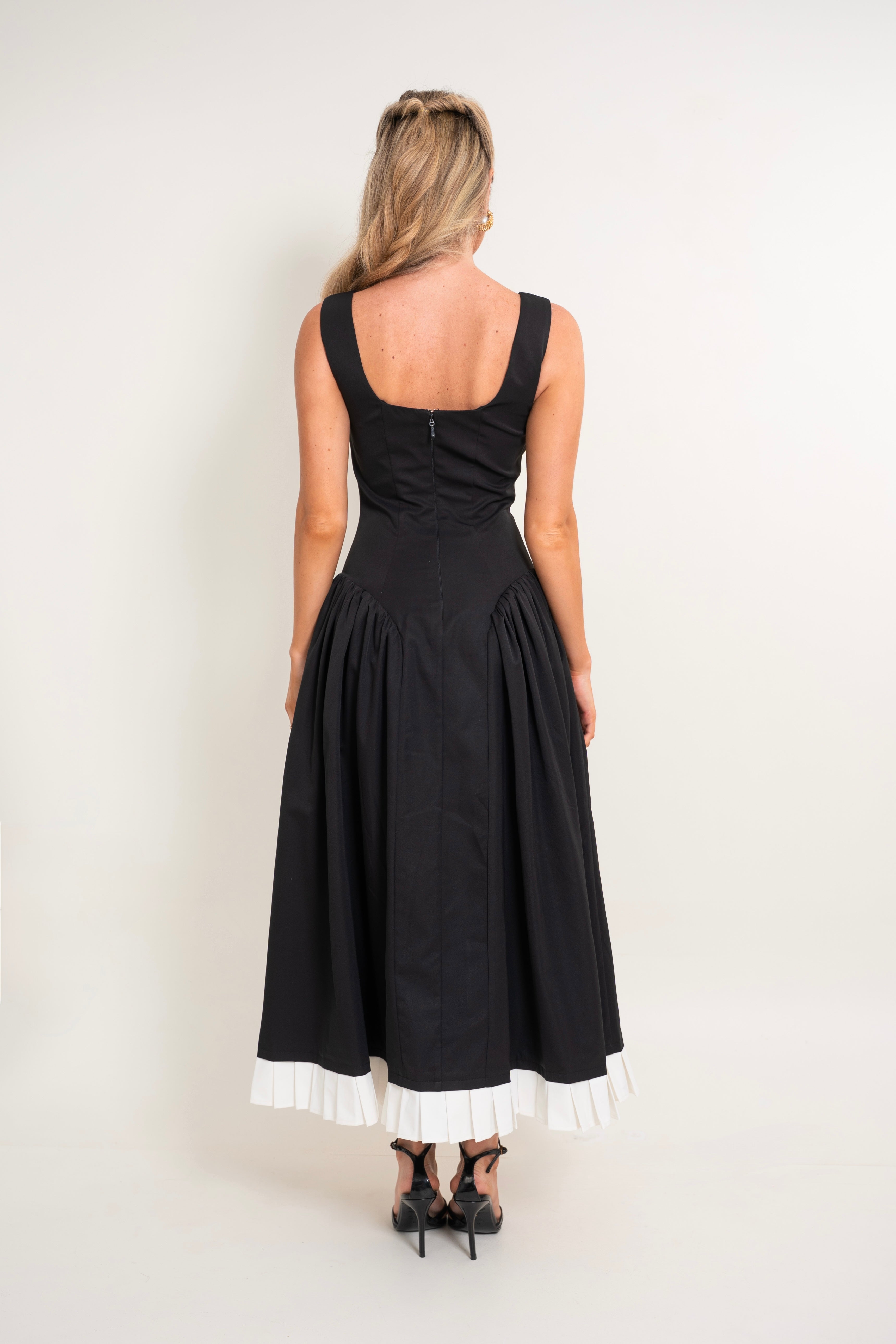 A woman viewed from the back wearing a sleeveless black midi dress with a gathered waist, pleated skirt, and contrasting white ruffled trim at the hem, paired with black heels against a plain light background.